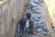 Police members removing a body covered on a stretcher from litter-strewn stormwater drain.