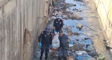 Police members removing a body covered on a stretcher from litter-strewn stormwater drain.