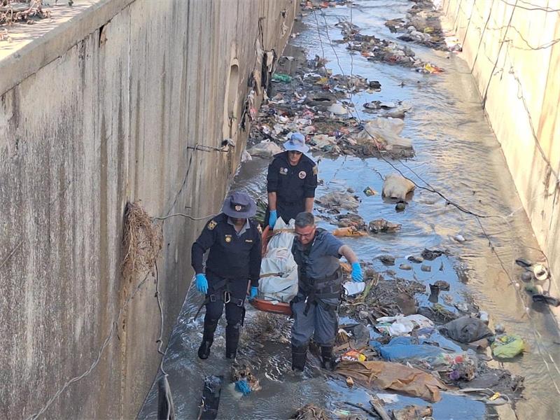 Police members removing a body covered on a stretcher from litter-strewn stormwater drain.