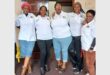 Five black women standing in a row all wearing white shirts, two in jeans and others in a black pants and a brown pants.
