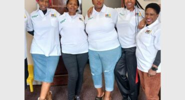 Five black women standing in a row all wearing white shirts, two in jeans and others in a black pants and a brown pants.