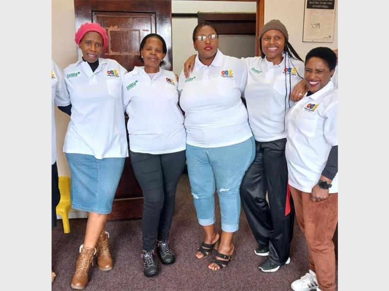 Five black women standing in a row all wearing white shirts, two in jeans and others in a black pants and a brown pants.