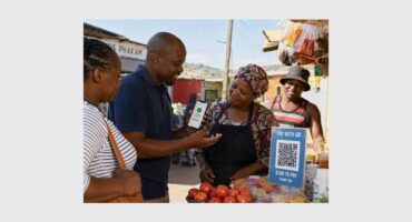 A black man speaking to three black females in a spaza shop setting while demonstrating a payment method.
