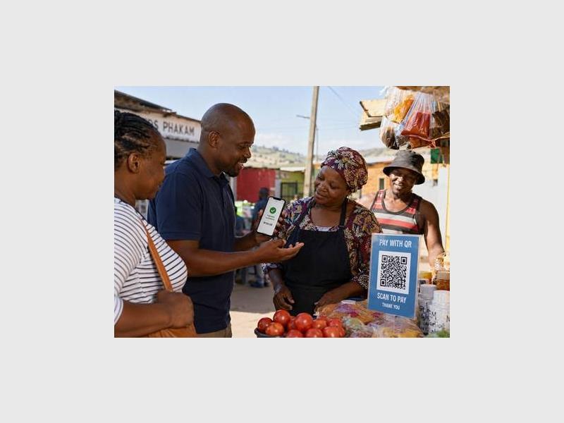 A black man speaking to three black females in a spaza shop setting while demonstrating a payment method.