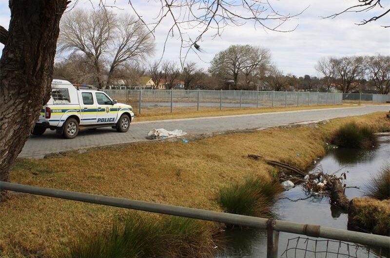 Photo of a marked SAPS vehicle standing next to a spruit with a body bag on the side