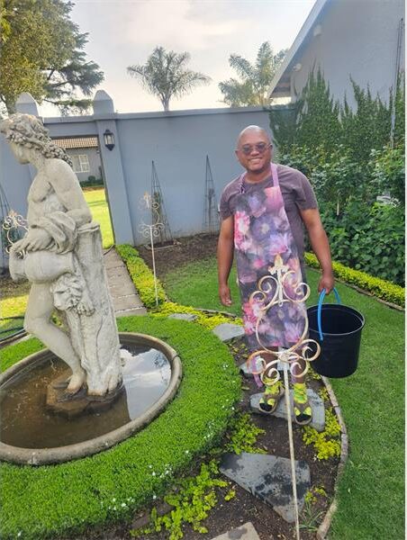 Coloured man in a floral apron holding a black bucket standing next to a water feature