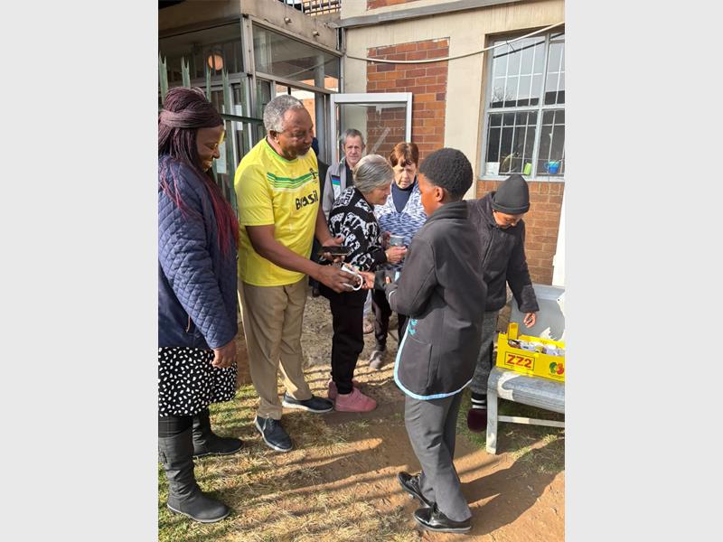 A young boy in school uniform hands a parcel over to a man in a yellow shirt while a woman looks on.