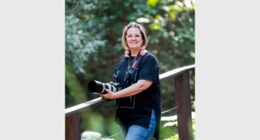 A white woman with blonde hair standing with her hands leaning on a wooden railing, wearing jeans and with a camera around her neck.