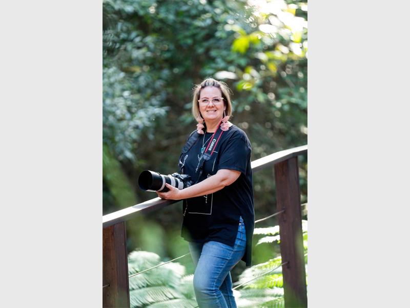 A white woman with blonde hair standing with her hands leaning on a wooden railing, wearing jeans and with a camera around her neck.
