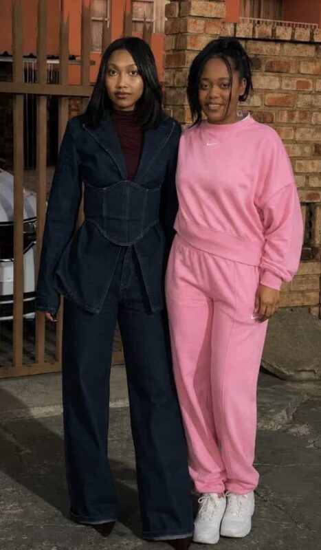 Two women standing at a palisade fence, one wearing all black and the other all pink.