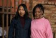 Two women standing at a palisade fence, one wearing all black and the other all pink.