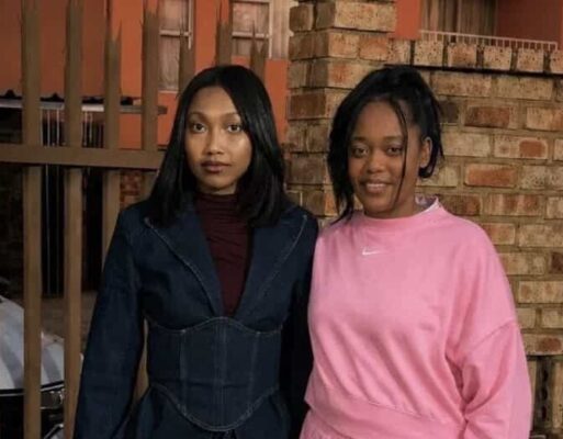 Two women standing at a palisade fence, one wearing all black and the other all pink.