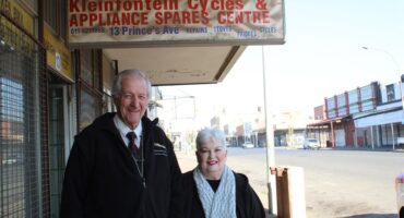 An elderly man and women stand next to a street under a weather-worn sign