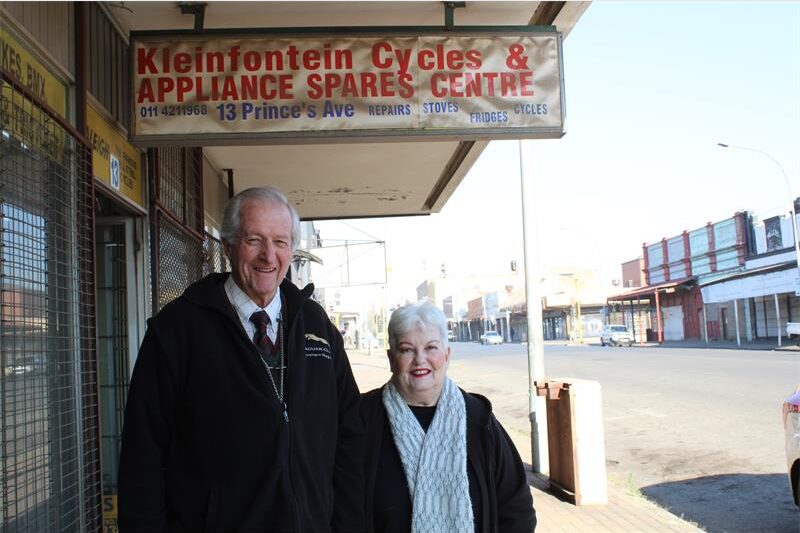 An elderly man and women stand next to a street under a weather-worn sign