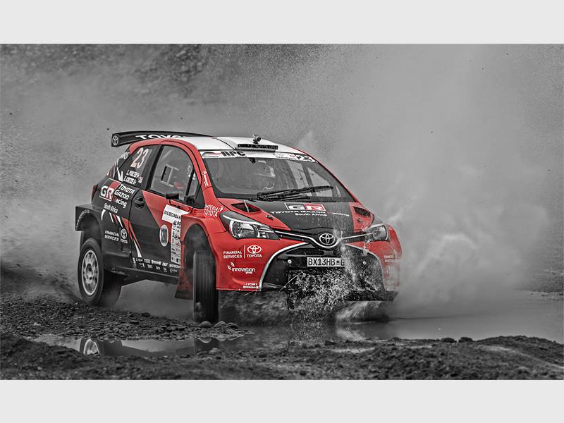 A red and black motorsport car in a race splashing up water and mud.