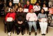 Five women standing in a row at the back of three women and a man seated, holding certificates.