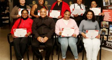Five women standing in a row at the back of three women and a man seated, holding certificates.
