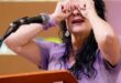 A woman standing behind a podium with a microphone on it holding her hands up to her eyes like binoculars.
