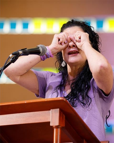 A woman standing behind a podium with a microphone on it holding her hands up to her eyes like binoculars.