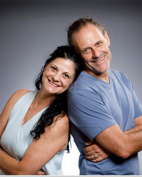A white woman and man standing back to back and smiling into the camera.