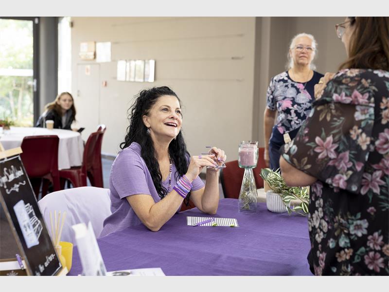 A woman with long dark hair sitting at a table with a purple tablecloth and speaking to other woman standing in front of her.