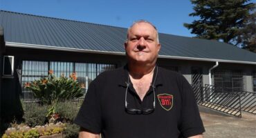 A white male wearing a black shirt with glasses around his neck standing in front of a building.