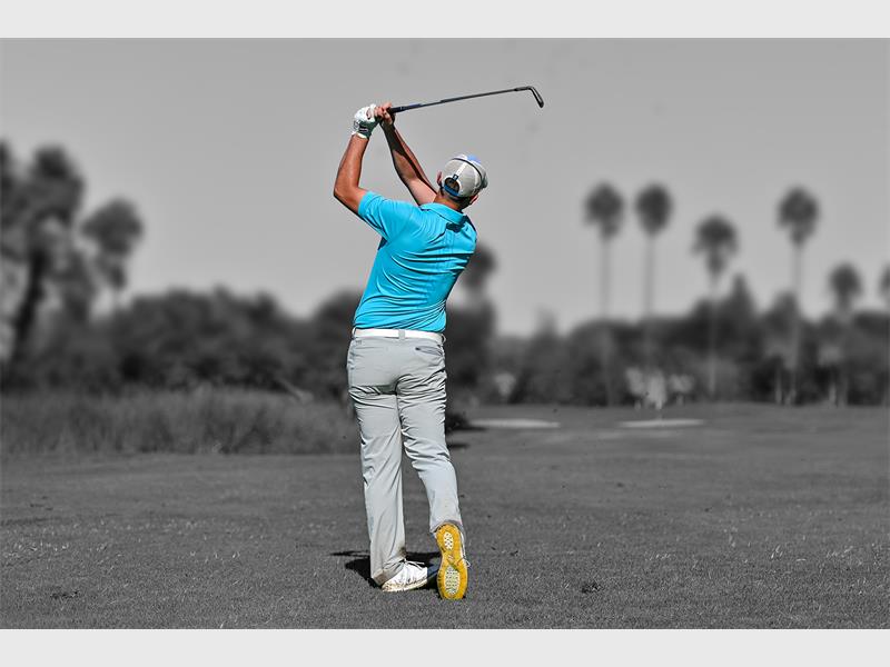 A photo of a golfer taken from behind as he swings and the golfer is in colour of white pants and blue shirt and the background in black and white.
