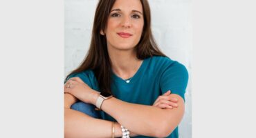 A white woman with long dark hair wearing a blue shirt and folding her arms over her chest.
