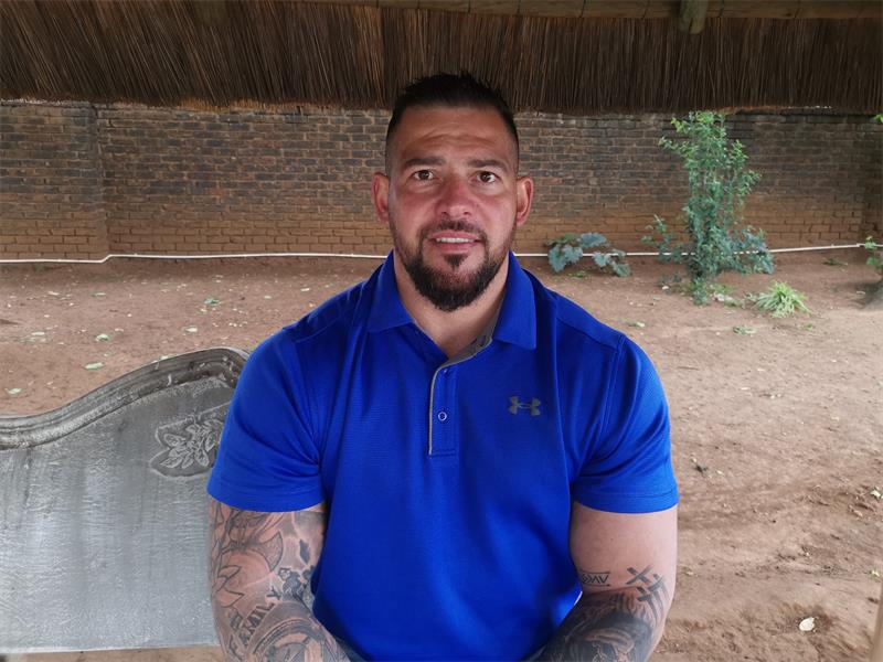 A white man with short dark hair, a beard and moustache and tatoos on both arms, wearing a blue shirt.