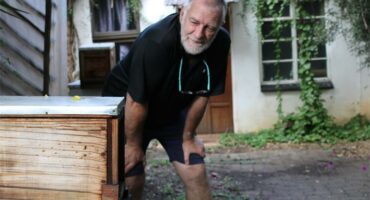 A white man with grey hair and beard wearing a black T-shirt with a pair of reading glasses dangling from his neck bending down with his hands on his knees next to a beehive.