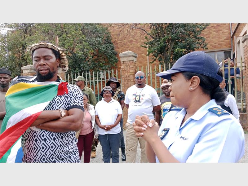 A black man dressed in zulu headband and wearing a south african flag over his chest standing with a police woman in uniform with people in the background outside a court.