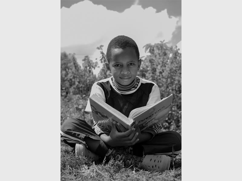 A black and white photo of a young black child reading a book to the background of an open field and sky.