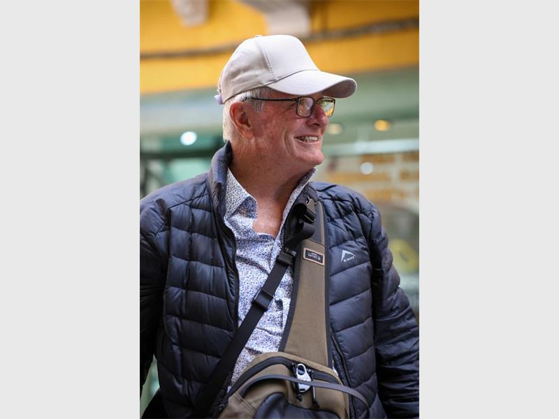 A white man looking away from the camera wearing a white cap, black jacket and bag over his chest.