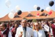 Man in a white suite standing next to a woman in a white blouse. Learners with red school uniform surrounding them.