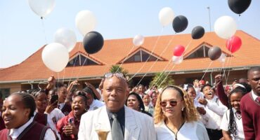 Man in a white suite standing next to a woman in a white blouse. Learners with red school uniform surrounding them.