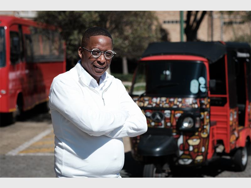 A black male standing in front of a red vehicle wearing a white shirt and glasses with his arms crossed over his chest.