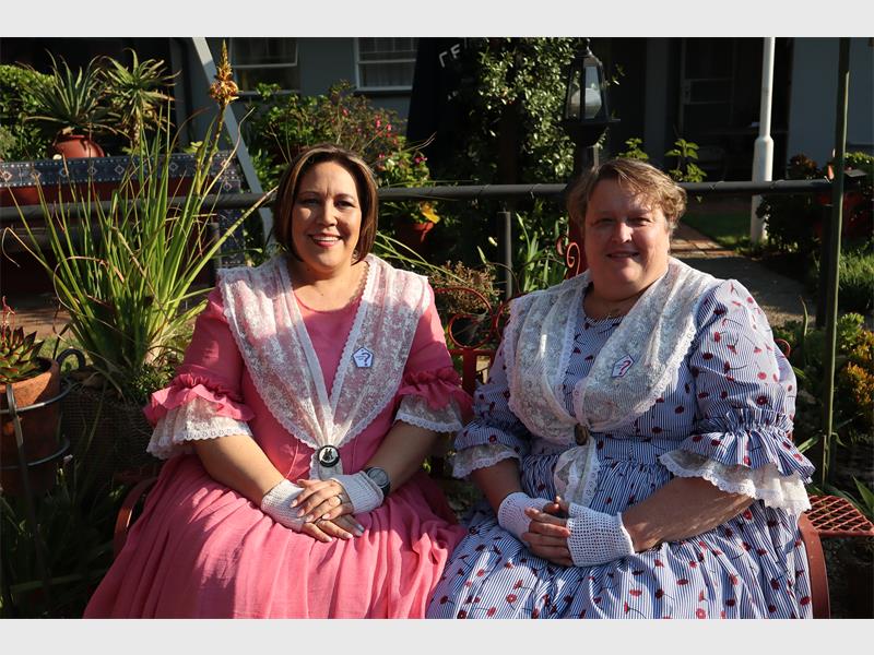 Two ladies sitting on a bench, one in a pink dress and one in a blue dress.