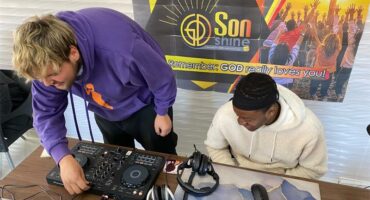 A white male and a black male seated behind a desk working on a laptop and looking at sound equipment.