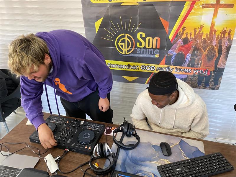A white male and a black male seated behind a desk working on a laptop and looking at sound equipment.