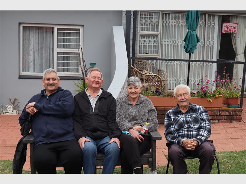 from left two elderly men sitting on bench, along with a lady and a man sitting on a chair