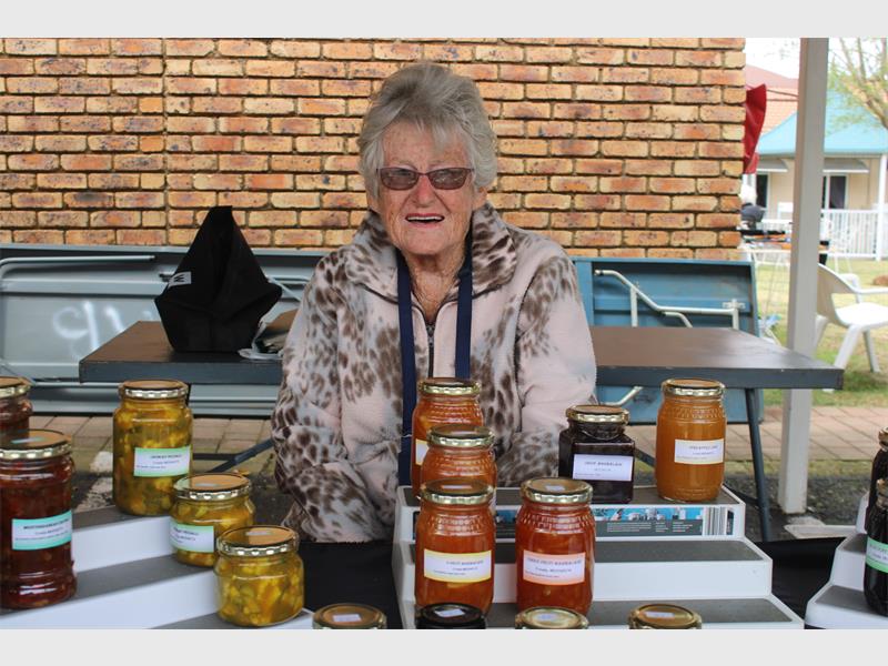 An elderly woman sitting behind a table with displays of jam on it.