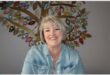 A white woman with grey hair wearing a light blue shirt, smiling at the camera with an artwork behind her on the wall.