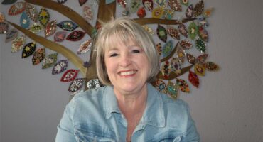 A white woman with grey hair wearing a light blue shirt, smiling at the camera with an artwork behind her on the wall.