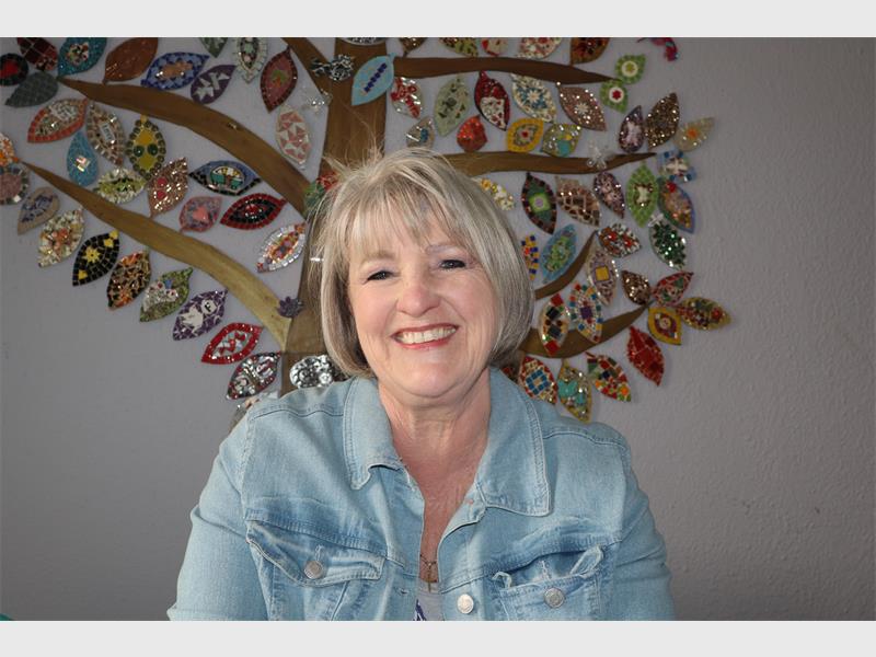A white woman with grey hair wearing a light blue shirt, smiling at the camera with an artwork behind her on the wall.