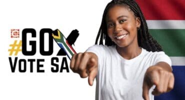 A young black female with dreadlocks pointing into the camera wearing a white T-shirt with a cross and the words go vote SA and the SA flag colours next to her.