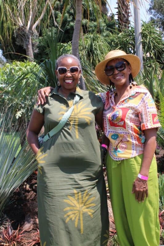 Two black women stand smiling with cycads in the background