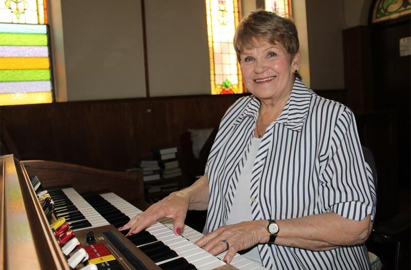 A woman smiles while sitting at an organ, her hand on the keys