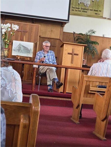 elderly white man sitting on a chair holding a mic and addressing a church congregation