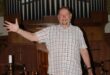 White man standing in front of a church with his right hand extended into the air