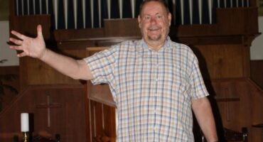 White man standing in front of a church with his right hand extended into the air